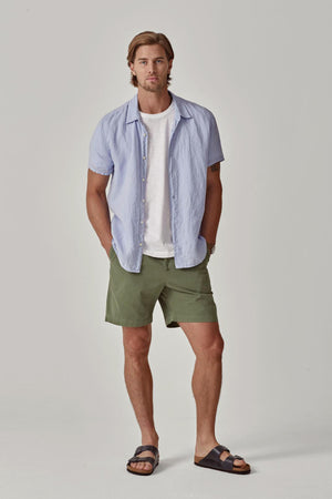 A man with shoulder-length hair stands wearing a Velvet by Graham & Spencer MACKIE LINEN BUTTON-UP SHIRT over a white t-shirt, green shorts, and black sandals, hands in pockets.