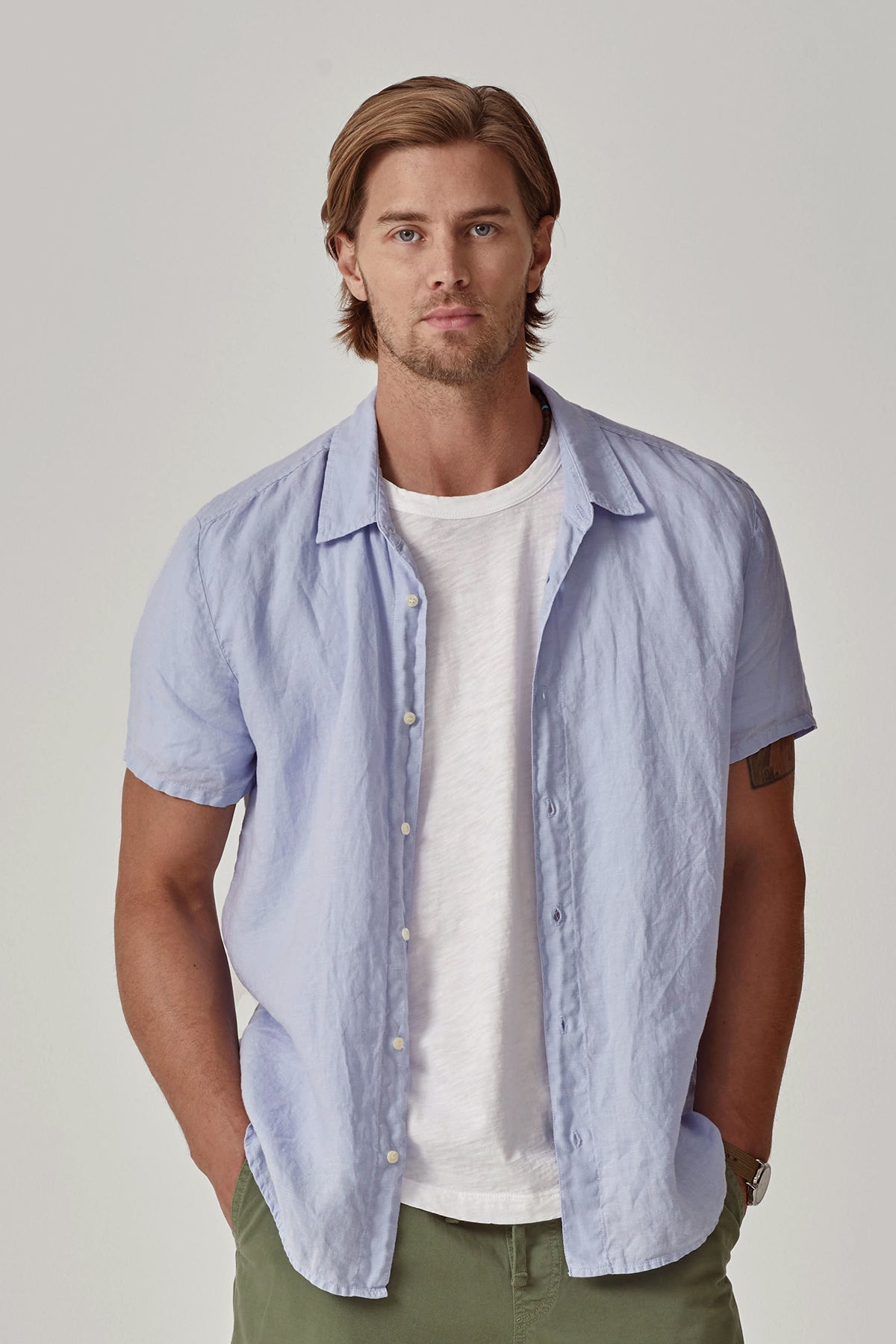   A man with short hair wears the MACKIE LINEN BUTTON-UP SHIRT by Velvet by Graham & Spencer in light blue over a white t-shirt, standing against a plain backdrop. The relaxed fit of his woven linen top enhances the effortlessly casual look. 