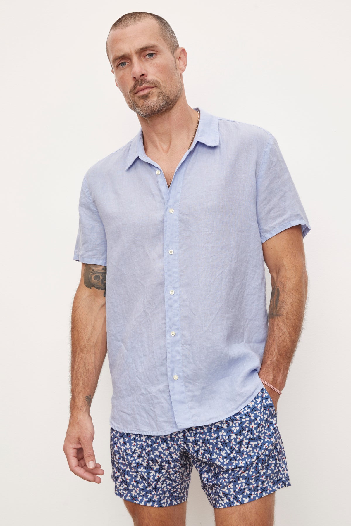   Man standing in a Velvet by Graham & Spencer MACKIE LINEN BUTTON-UP SHIRT and patterned shorts, looking at the camera with a neutral expression. 