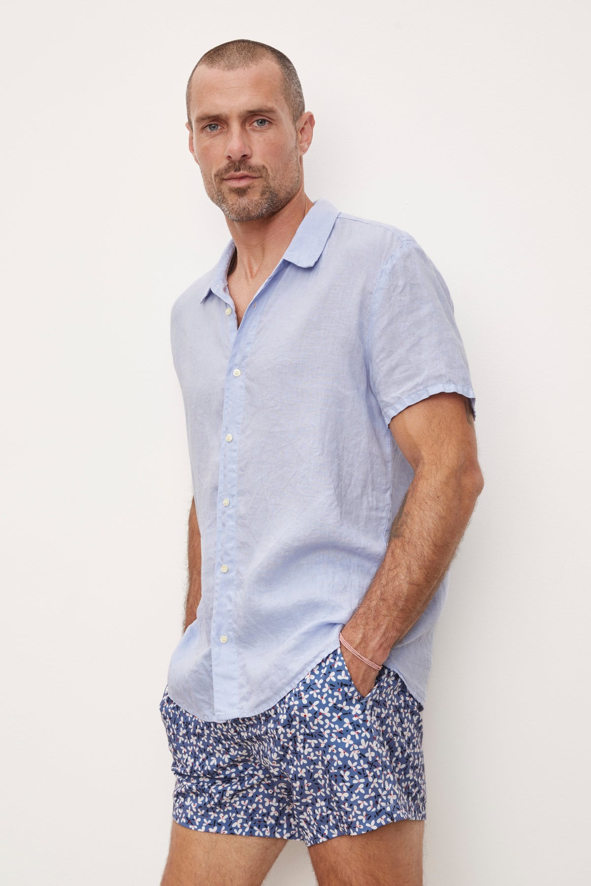   A man in a MACKIE LINEN BUTTON-UP SHIRT by Velvet by Graham & Spencer and patterned shorts standing against a white background, hands in pockets, looking at the camera. 