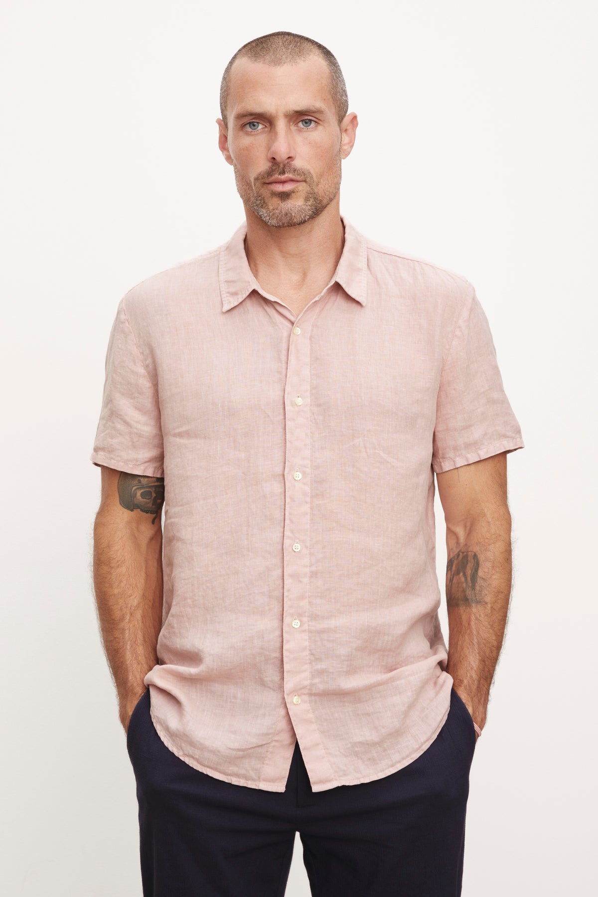   A man with a beard stands wearing a Velvet by Graham & Spencer MACKIE LINEN BUTTON-UP SHIRT in pink and navy pants, looking directly at the camera. He has tattoos visible on his arms. 