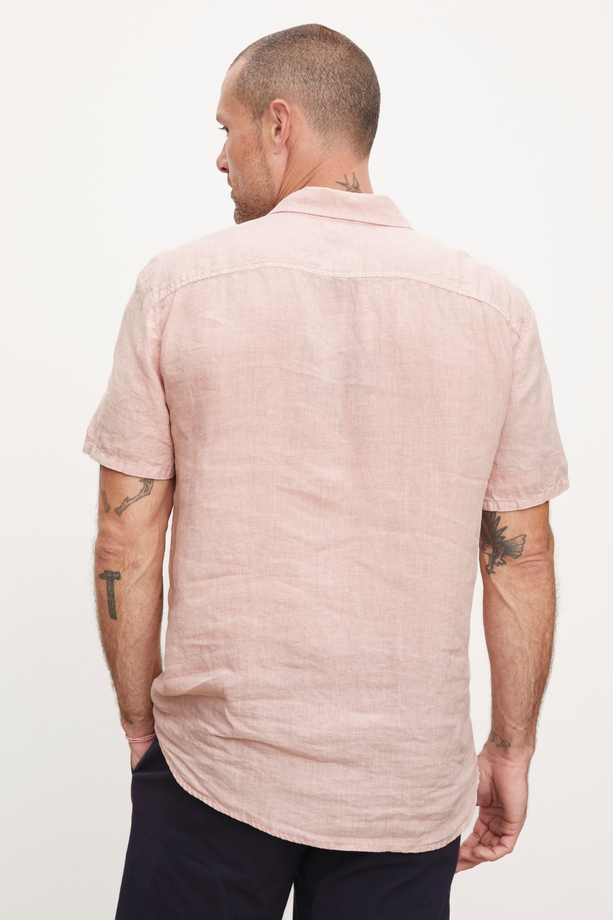 A man viewed from behind, wearing a Velvet by Graham & Spencer MACKIE LINEN BUTTON-UP SHIRT and dark pants, displaying visible tattoos on his arms.-36753539694785
