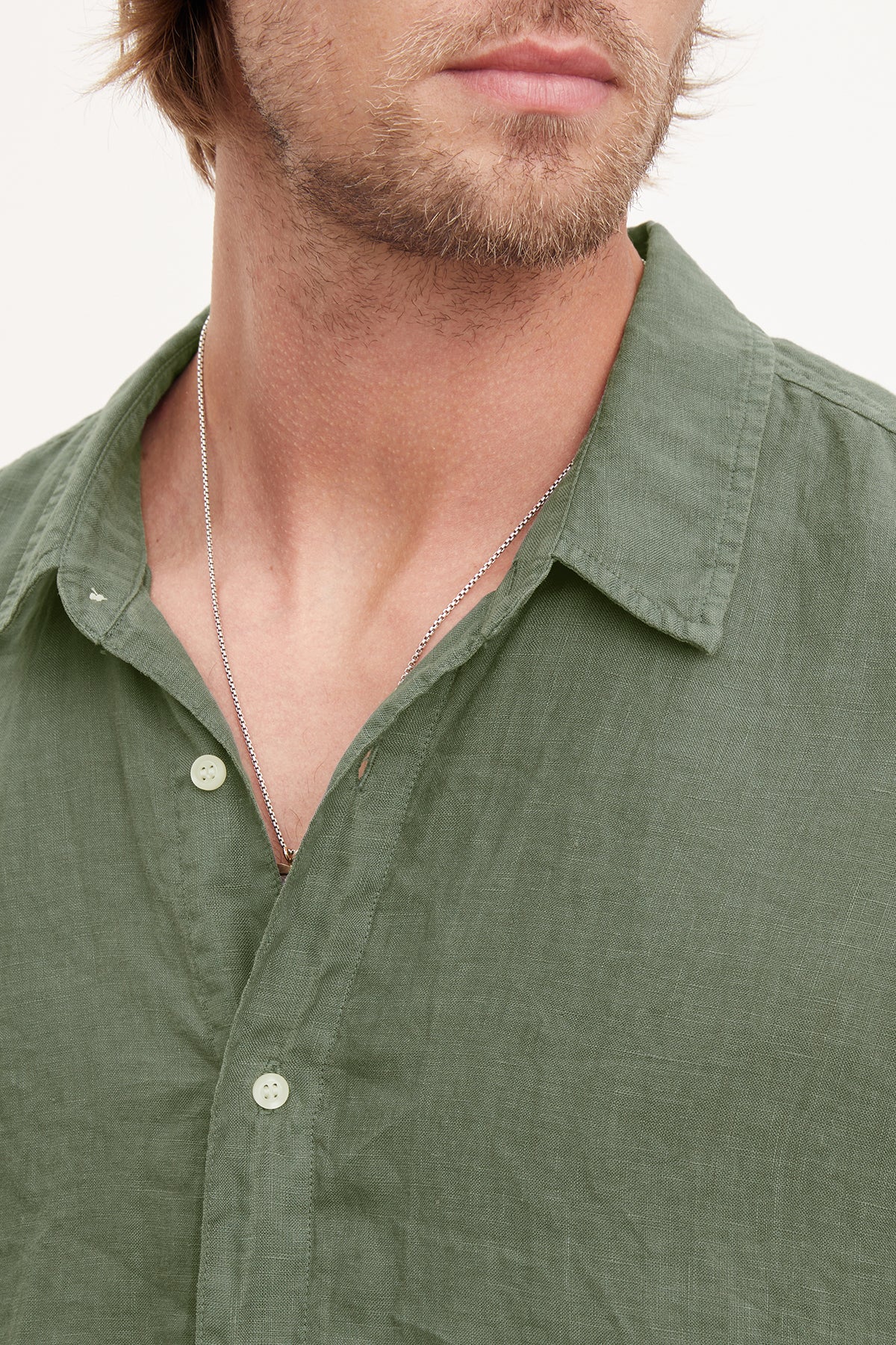   Close-up of a person with a beard wearing an olive green MACKIE LINEN BUTTON-UP SHIRT by Velvet by Graham & Spencer and a silver necklace. The image focuses on the short sleeve shirt's collar and upper chest area, perfect for a summer wardrobe. 