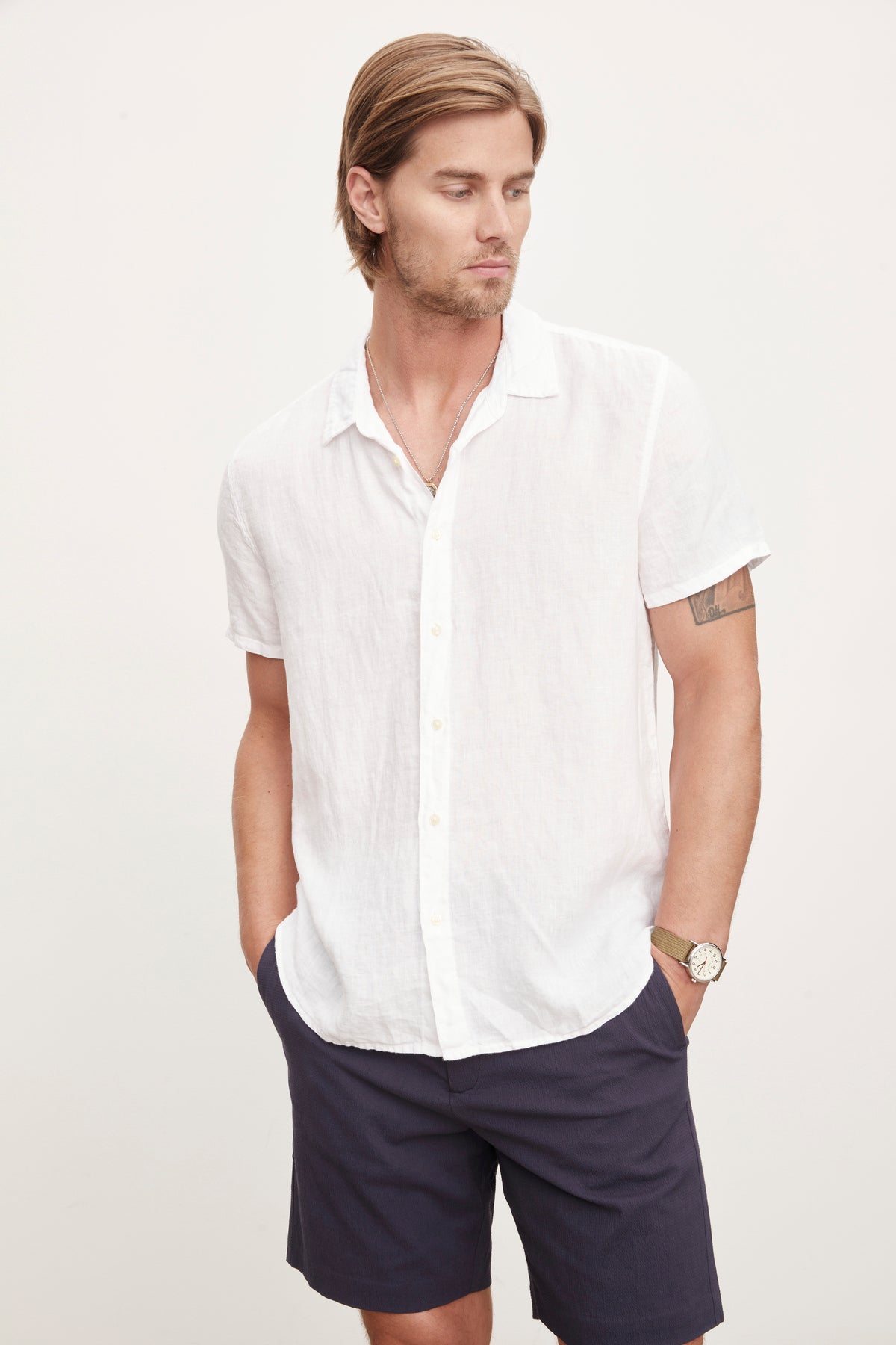 A man in a Velvet by Graham & Spencer MACKIE LINEN BUTTON-UP SHIRT and navy shorts, standing with one hand in his pocket and a watch on his left wrist, epitomizes the relaxed fit of a summer wardrobe.-36753541267649