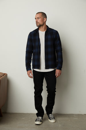 A man wearing a Velvet by Graham & Spencer MARK ZIP-UP PLAID SHIRT and black pants in front of a wall.