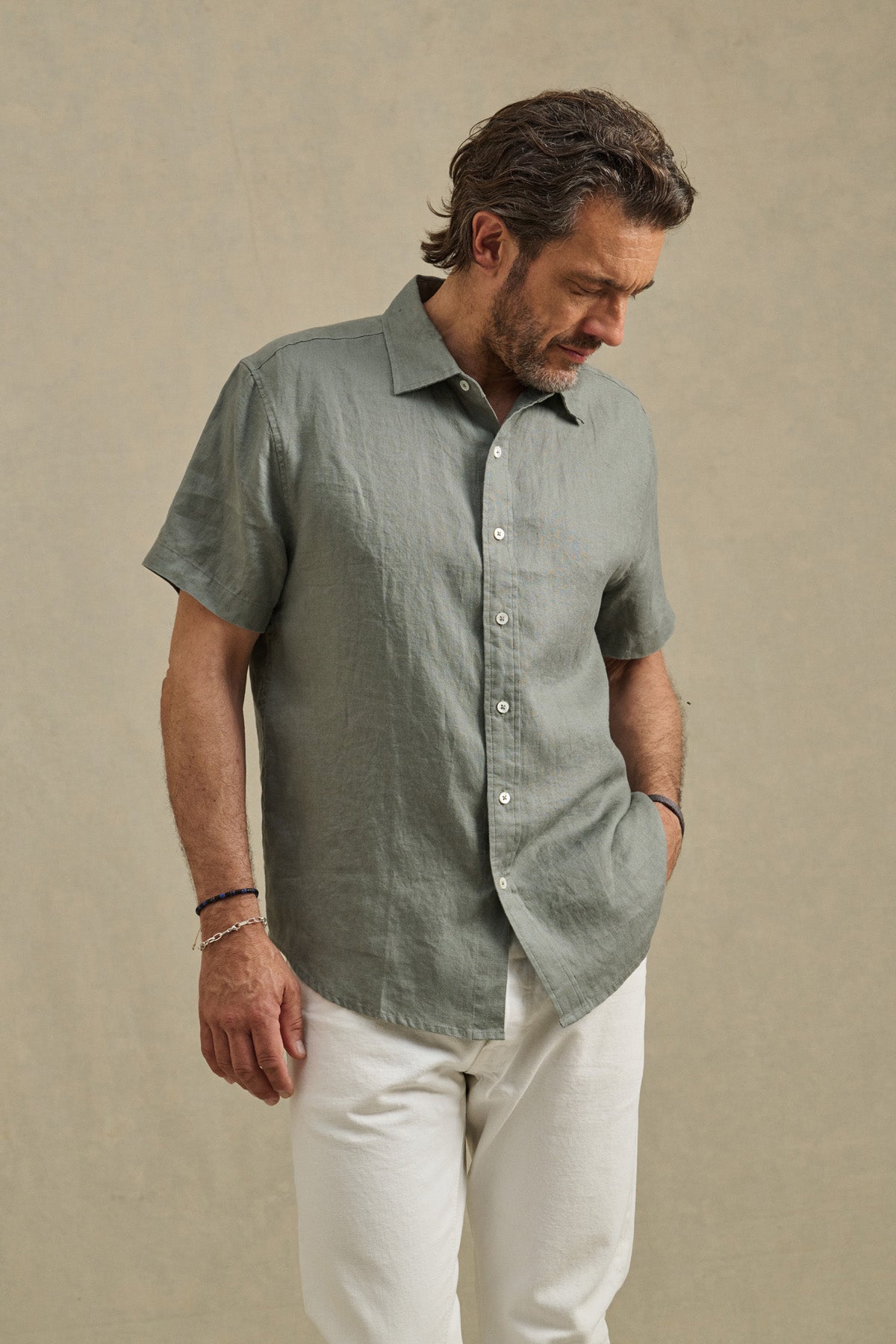 A man wears the MICAH LINEN SHIRT by Velvet by Graham & Spencer—a light green textured button-up—paired with white pants, standing against a neutral background and looking down with one hand at his side.-41479923335361