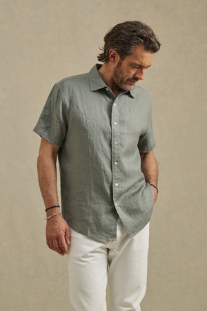 A man wears the MICAH LINEN SHIRT by Velvet by Graham & Spencer—a light green textured button-up—paired with white pants, standing against a neutral background and looking down with one hand at his side.