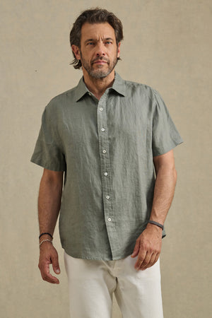 A man wears the MICAH LINEN SHIRT by Velvet by Graham & Spencer—a short-sleeve, light grey-green textured linen button-up—paired with white pants against a plain beige background.