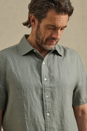 A man with wavy brown hair and a beard looks down while wearing the MICAH LINEN SHIRT, a light green short-sleeve button-up by Velvet by Graham & Spencer, crafted from breathable linen fabric.