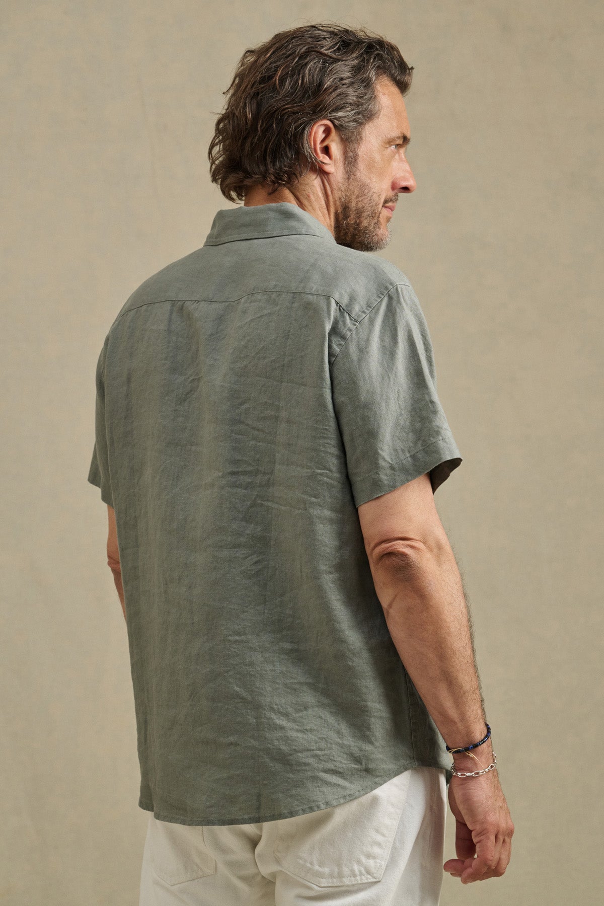 A man wears the Velvet by Graham & Spencer MICAH LINEN SHIRT, a short-sleeve, light gray-green textured linen button-up, with white pants, standing and facing away against a neutral background.
