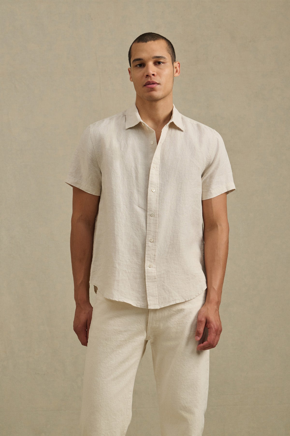 A man stands against a plain beige background, wearing the MICAH LINEN SHIRT by Velvet by Graham & Spencer—a short-sleeve, textured linen shirt—paired with matching light-colored breathable pants.