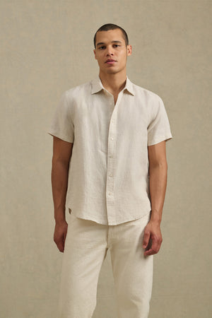 A man stands against a plain beige background, wearing the MICAH LINEN SHIRT by Velvet by Graham & Spencer—a short-sleeve, textured linen shirt—paired with matching light-colored breathable pants.