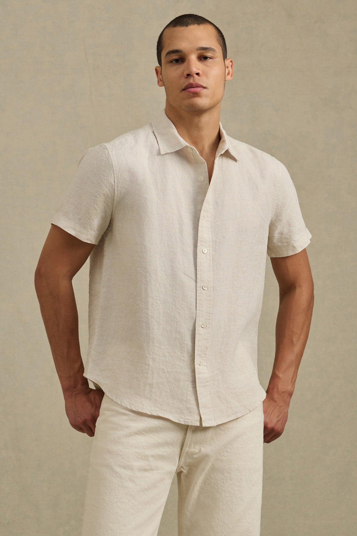 Man wearing the MICAH LINEN SHIRT by Velvet by Graham & Spencer— a short-sleeve, light beige textured linen shirt—paired with light-colored pants, standing against a neutral background.