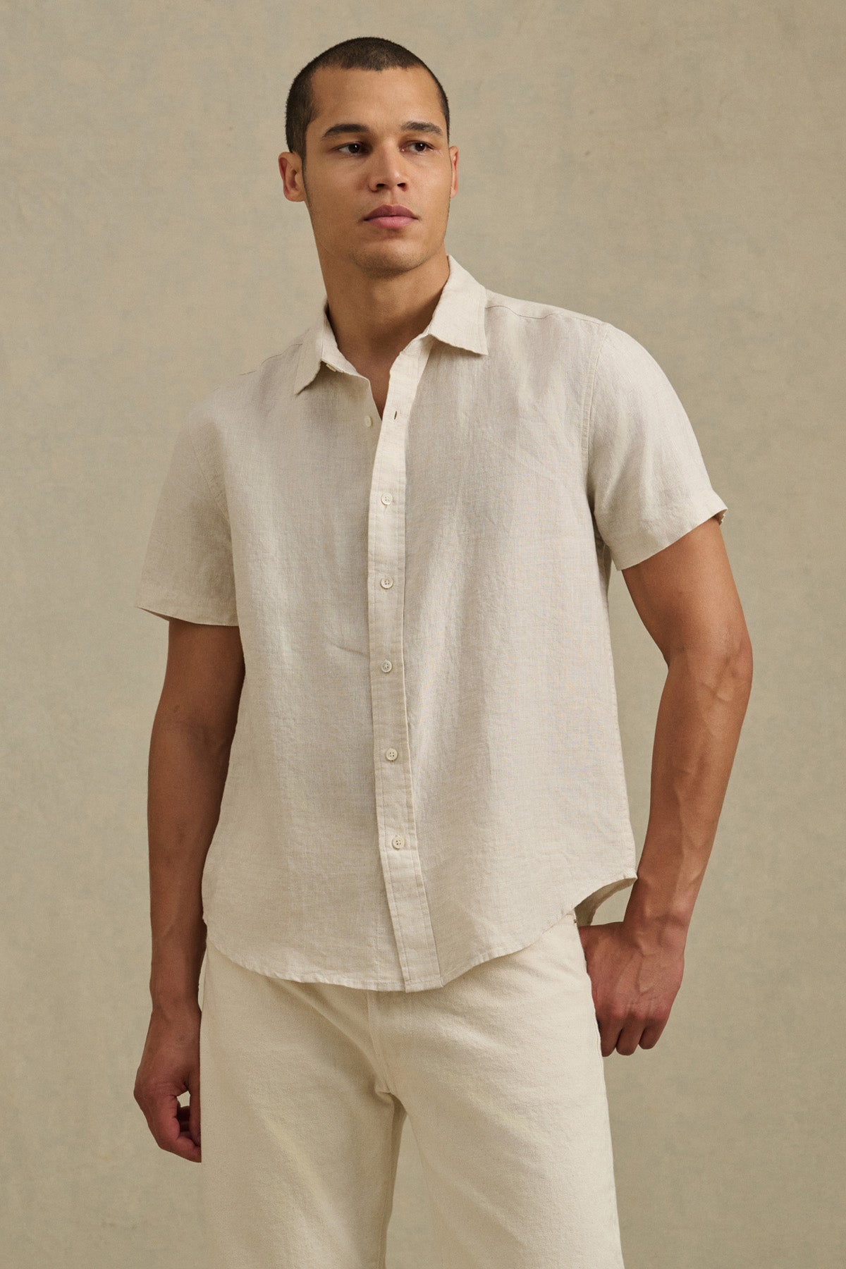 A man wears the MICAH LINEN SHIRT by Velvet by Graham & Spencer—a short-sleeve, light beige button-up made from breathable linen—with matching pants, standing against a plain, neutral background.-41374398415041