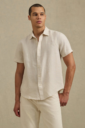 A man wears the MICAH LINEN SHIRT by Velvet by Graham & Spencer—a short-sleeve, light beige button-up made from breathable linen—with matching pants, standing against a plain, neutral background.