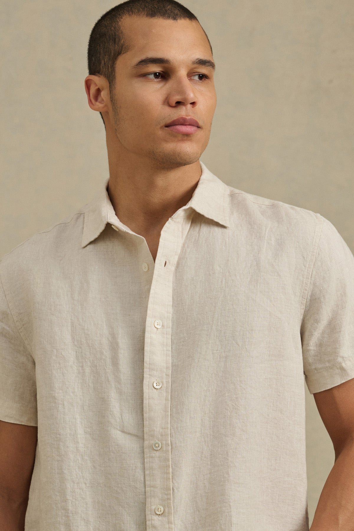A person models the MICAH LINEN SHIRT by Velvet by Graham & Spencer—a light beige, short-sleeve textured linen shirt—against a neutral background, gazing off to the side.