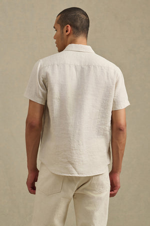 A man stands facing away, wearing the MICAH LINEN SHIRT by Velvet by Graham & Spencer—a short-sleeve, light beige textured linen shirt and matching breathable pants—set against a plain, neutral background.