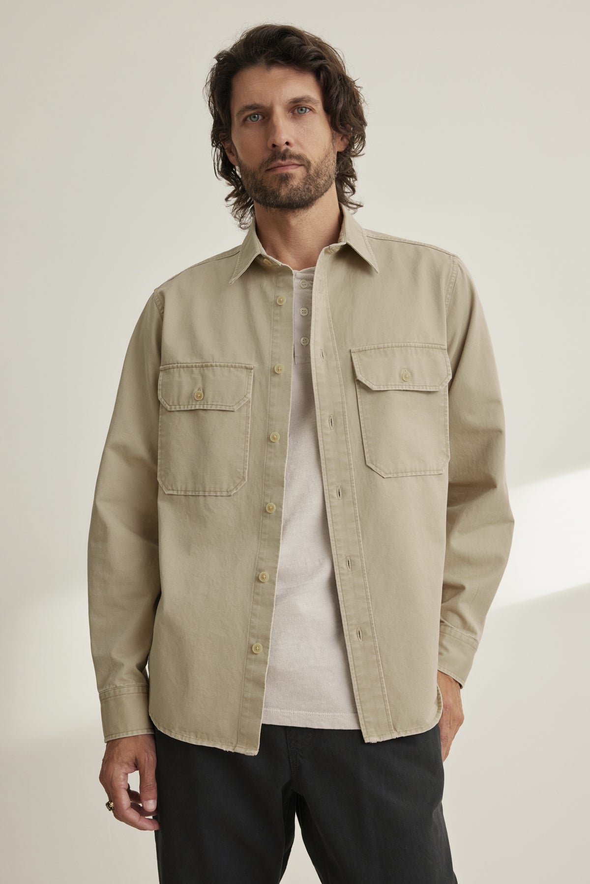 A man with wavy hair wears a Velvet by Graham & Spencer ORLANDO BUTTON-UP SHIRT in beige over a light shirt and black pants, standing in a softly lit room.-38643339919553