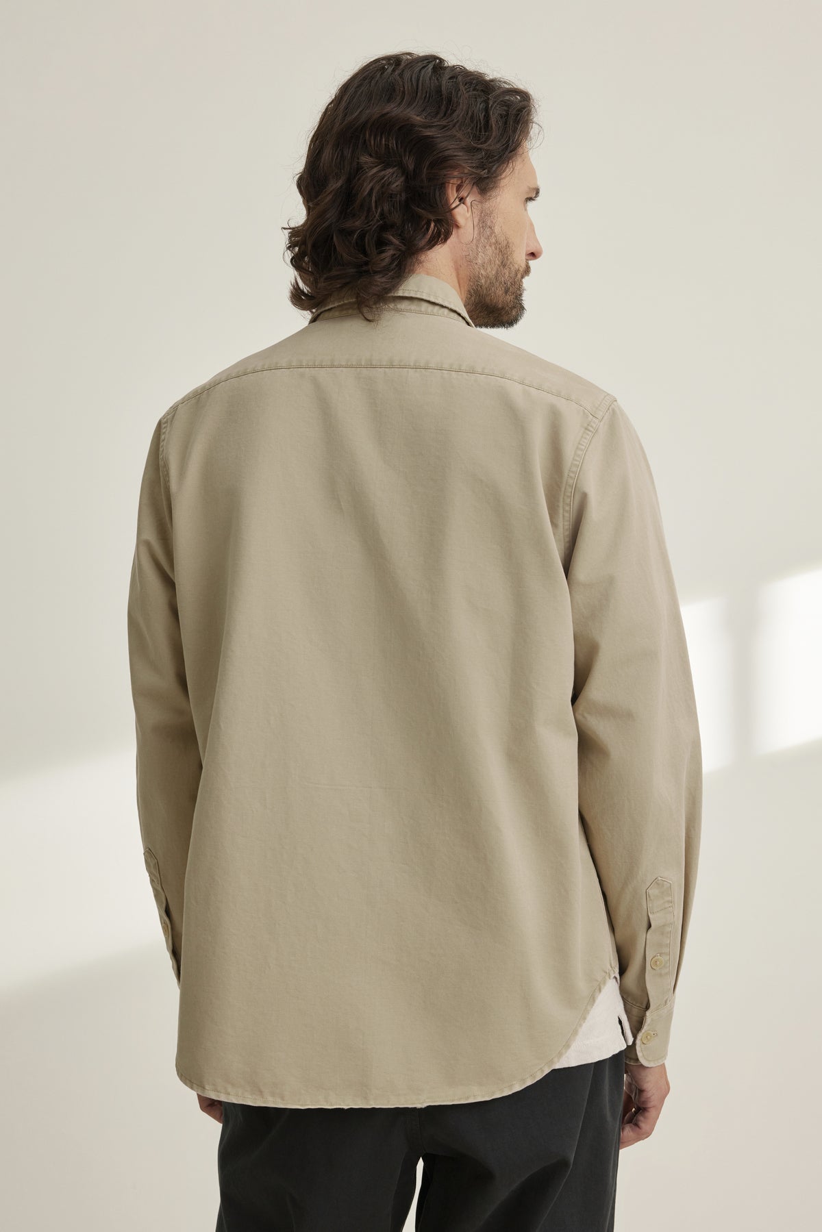 A person with medium-length hair is facing away, wearing an ORLANDO BUTTON-UP SHIRT by Velvet by Graham & Spencer and dark pants, standing in a softly lit room.-38643339854017
