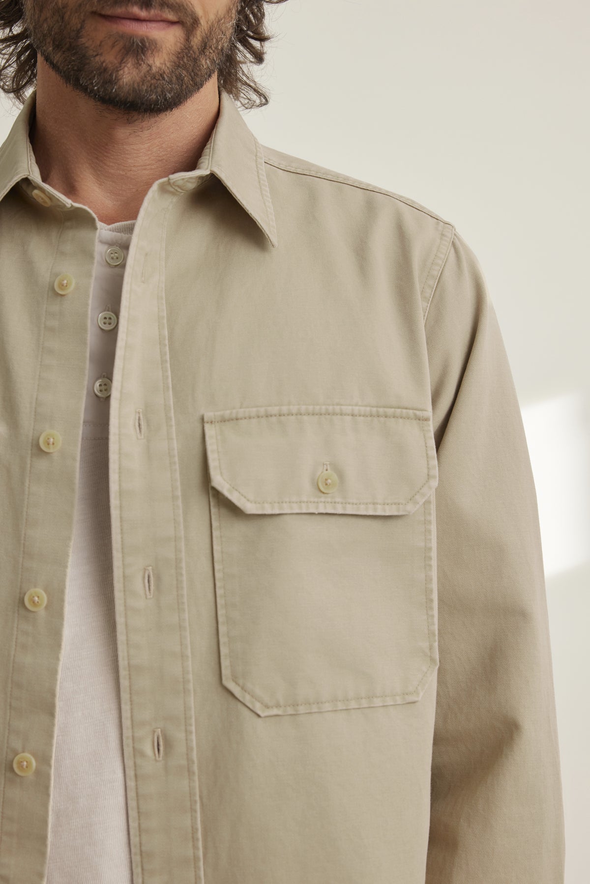 A bearded man partially appears against a light background, wearing a partially open ORLANDO BUTTON-UP SHIRT by Velvet by Graham & Spencer in beige, with a chest pocket, over a white undershirt.-38643339821249