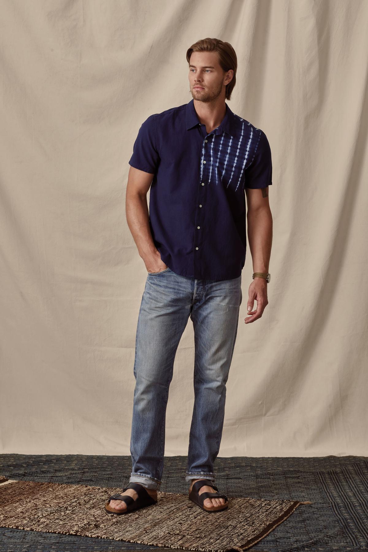 A man stands on a rug, wearing a Velvet by Graham & Spencer Rafael button-up shirt, jeans, and black sandals, with one hand slightly raised.-36753533763777