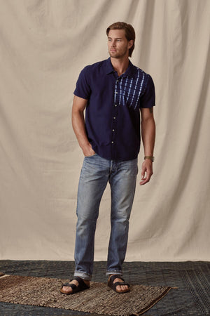 A man stands on a rug, wearing a Velvet by Graham & Spencer Rafael button-up shirt, jeans, and black sandals, with one hand slightly raised.
