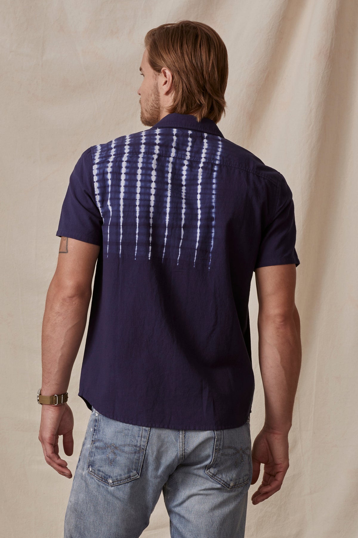 A man with long blonde hair stands with his back facing the camera, wearing a dark purple RAFAEL BUTTON-UP SHIRT by Velvet by Graham & Spencer with a white tie-dye design on the back and light blue jeans.-36753533829313
