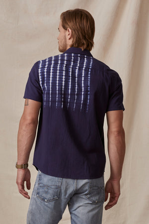 A man with long blonde hair stands with his back facing the camera, wearing a dark purple RAFAEL BUTTON-UP SHIRT by Velvet by Graham & Spencer with a white tie-dye design on the back and light blue jeans.