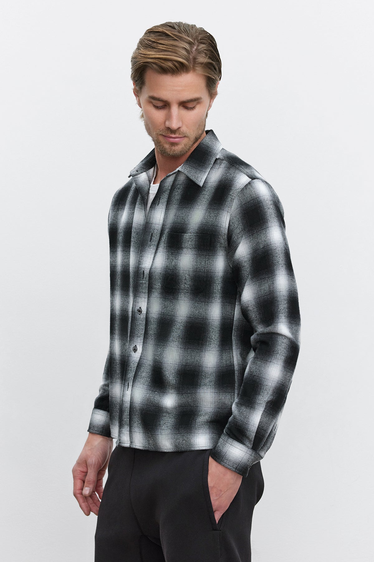 A man is looking down while wearing the STANTON BUTTON-UP SHIRT with a black and white plaid ombre design from Velvet by Graham & Spencer, along with dark pants against a plain background.-37893756453057