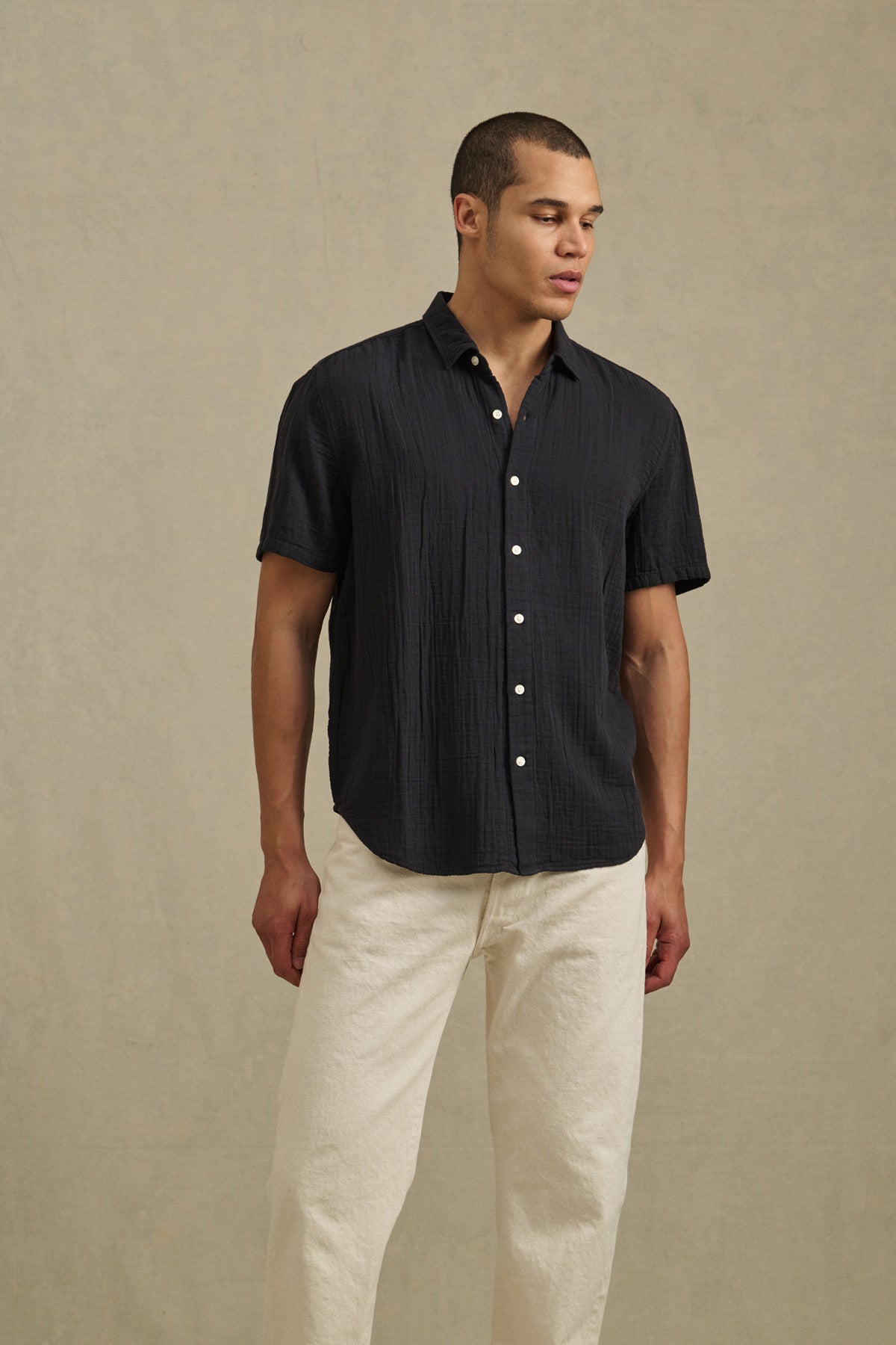 A man wears the TROY SHIRT by Velvet by Graham & Spencer— a short sleeve cotton gauze button-up—paired with off-white pants, standing against a plain, light-colored background.-41428172538049