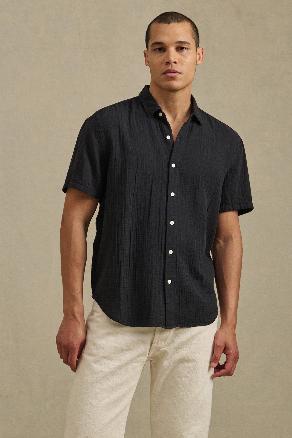 A man wears the TROY SHIRT by Velvet by Graham & Spencer— a short sleeve cotton gauze button-up—paired with light-colored pants, standing against a plain beige background.-41428172570817