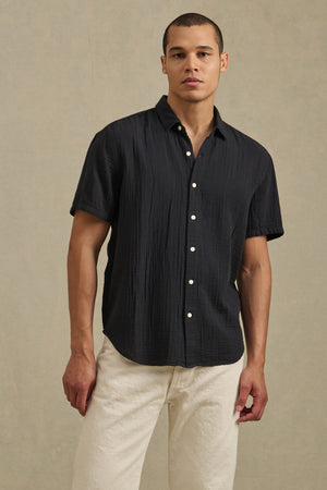 A man wears the TROY SHIRT by Velvet by Graham & Spencer— a short sleeve cotton gauze button-up—paired with light-colored pants, standing against a plain beige background.