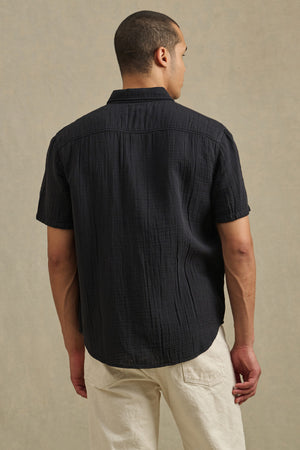 A person stands facing away, wearing the TROY SHIRT by Velvet by Graham & Spencer with light-colored pants, against a plain beige background.
