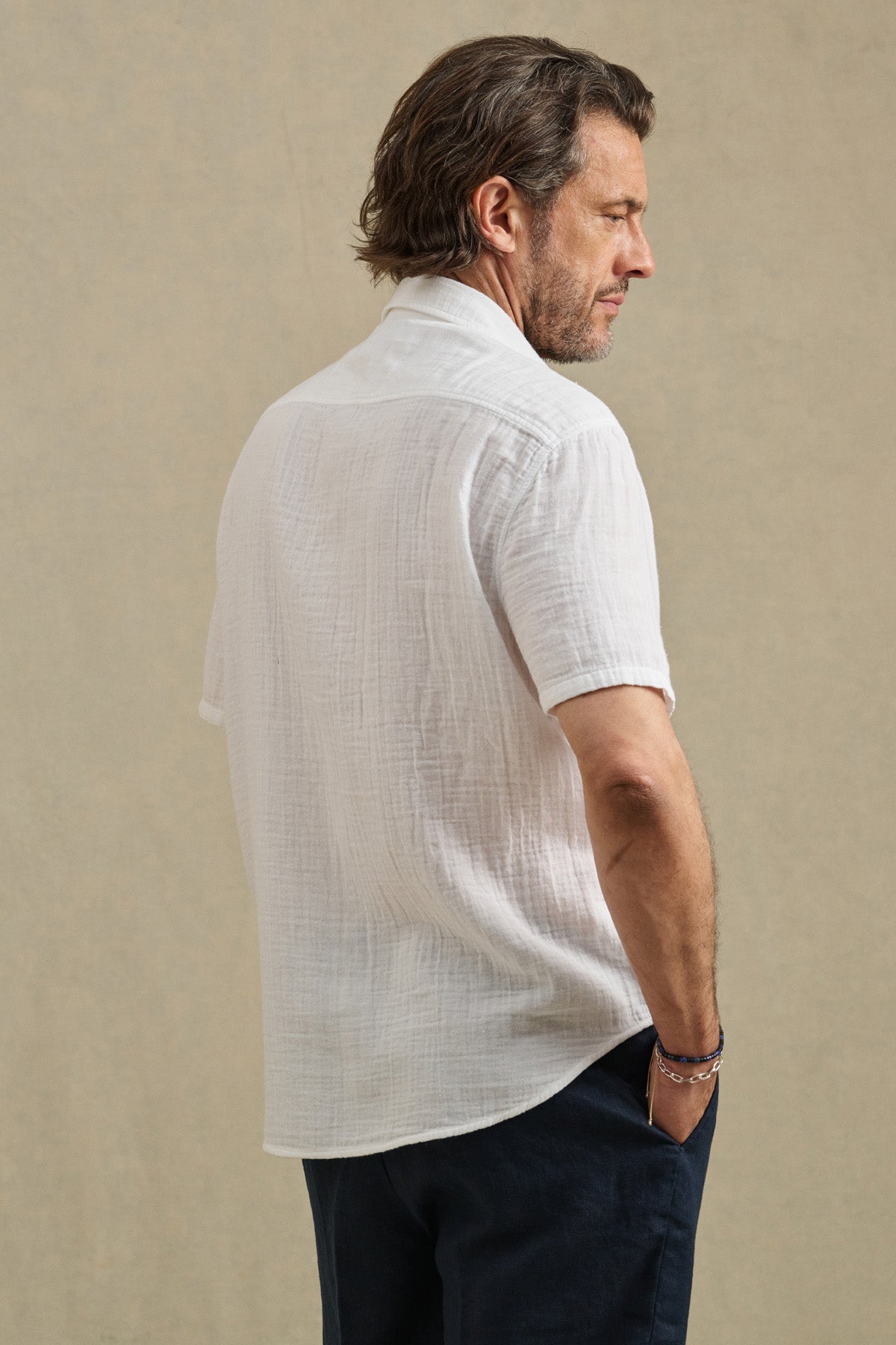 A man faces away, wearing the TROY SHIRT by Velvet by Graham & Spencer—a white cotton gauze button-up short sleeve shirt—with dark pants, set against a plain beige background.