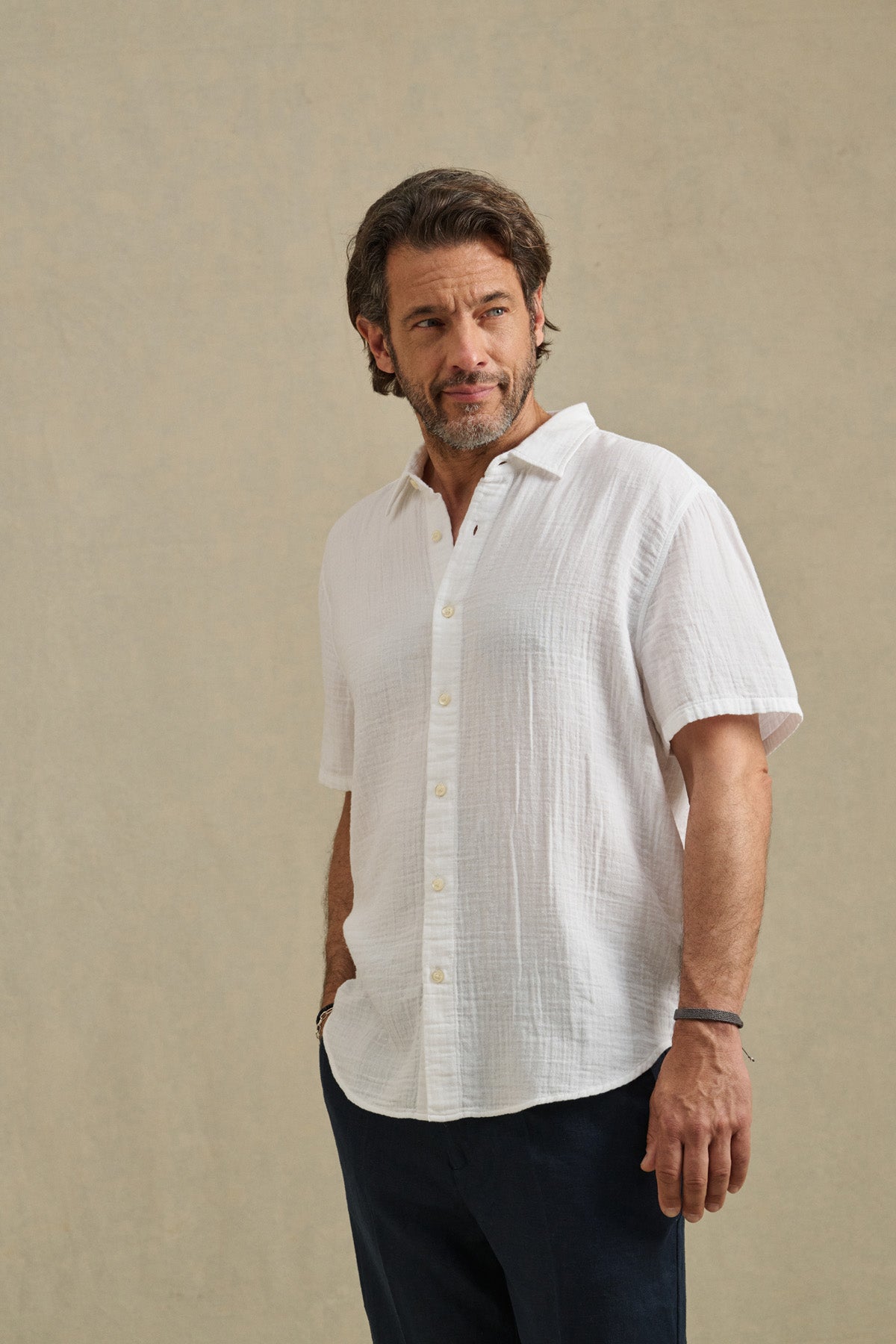 A man with short brown hair and a beard stands against a neutral background, wearing the TROY SHIRT by Velvet by Graham & Spencer—a white cotton gauze button-up short sleeve shirt—and dark pants, with one hand in his pocket.