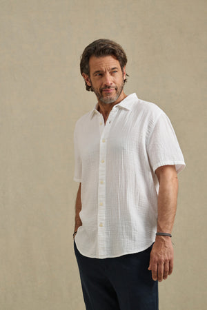 A man with short brown hair and a beard stands against a neutral background, wearing the TROY SHIRT by Velvet by Graham & Spencer—a white cotton gauze button-up short sleeve shirt—and dark pants, with one hand in his pocket.
