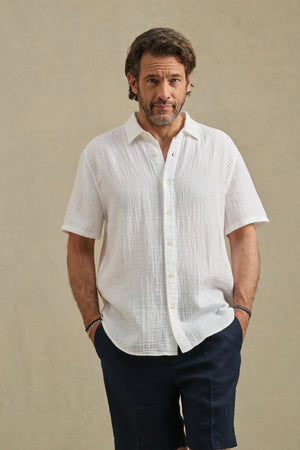 A man stands against a plain background, hands in pockets, wearing the TROY SHIRT from Velvet by Graham & Spencer— a white cotton gauze short-sleeve button-up—paired with dark shorts.
