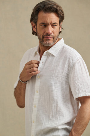 A man with medium-length hair and a beard wears the TROY SHIRT by Velvet by Graham & Spencer, a white cotton gauze short sleeve button-up, looking to the side against a plain, neutral background.