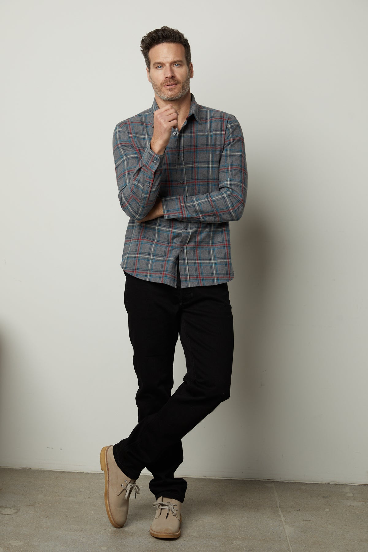   A man wearing the WILDER PLAID BUTTON-UP SHIRT from Velvet by Graham & Spencer, crafted from cotton woven fabric, stands against a plain white wall, resting one hand on his chin and the other in his pocket. 