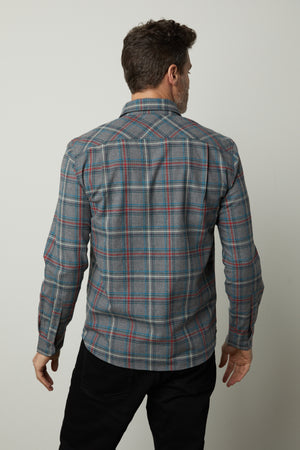 A man is standing with his back to the camera, showcasing the WILDER PLAID BUTTON-UP SHIRT by Velvet by Graham & Spencer, which features a grey and blue plaid pattern and is made from lightweight cotton. He pairs the shirt with black pants against a plain white background.