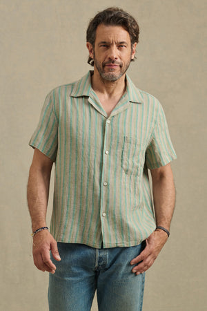 A man wears the WORTH SHIRT by Velvet by Graham & Spencer—a striped cotton-linen camp collar shirt—paired with blue jeans, standing against a plain beige background.
