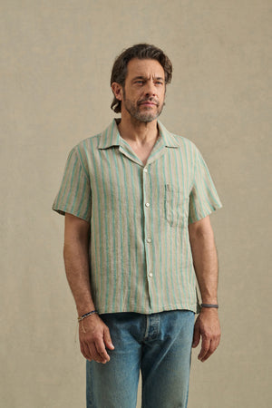 A man with short brown hair and a beard wears the WORTH SHIRT by Velvet by Graham & Spencer—a camp collar light green and beige striped cotton-linen blend shirt—paired with blue jeans, standing against a plain beige background.