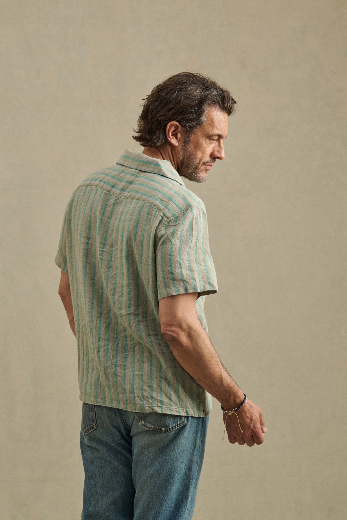 A man, seen from behind against a plain beige background, wears Velvet by Graham & Spencer's WORTH SHIRT—a short-sleeve, light green striped camp-collar shirt—paired with blue jeans.