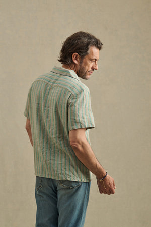 A man, seen from behind against a plain beige background, wears Velvet by Graham & Spencer's WORTH SHIRT—a short-sleeve, light green striped camp-collar shirt—paired with blue jeans.