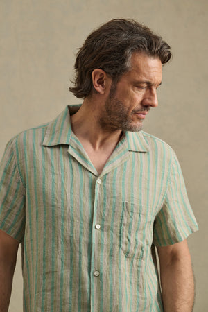 A man with medium-length brown hair and a beard wears the Velvet by Graham & Spencer WORTH SHIRT—a short-sleeved, striped cotton-linen blend camp collar shirt—looking down to his right against a neutral background.