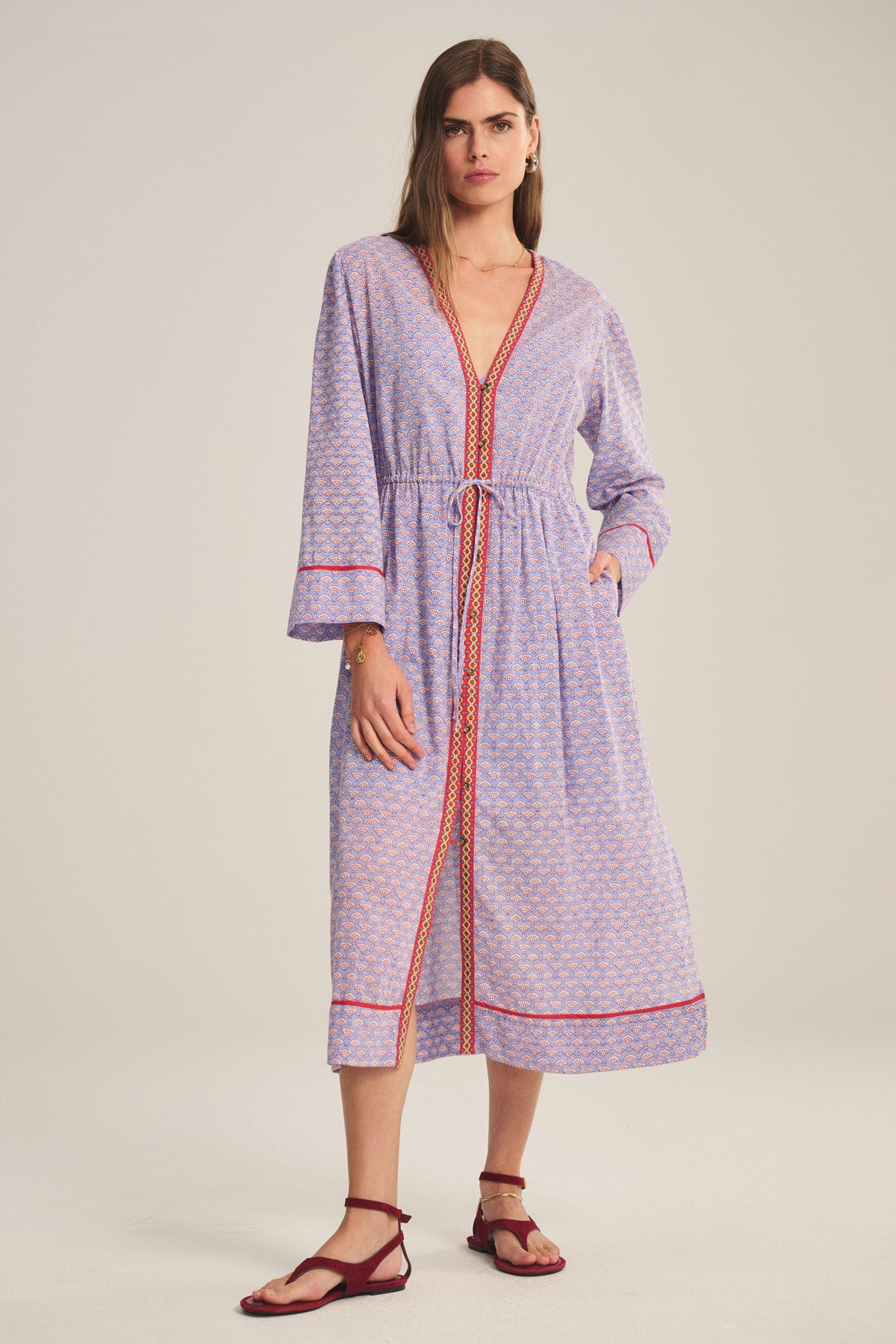 A woman wears the Velvet by Graham & Spencer WRENLEY DRESS, a long-sleeved, light purple patterned midi with red trim, tie waist, and embroidery, styled with flat red sandals against a plain background.-40808771453121