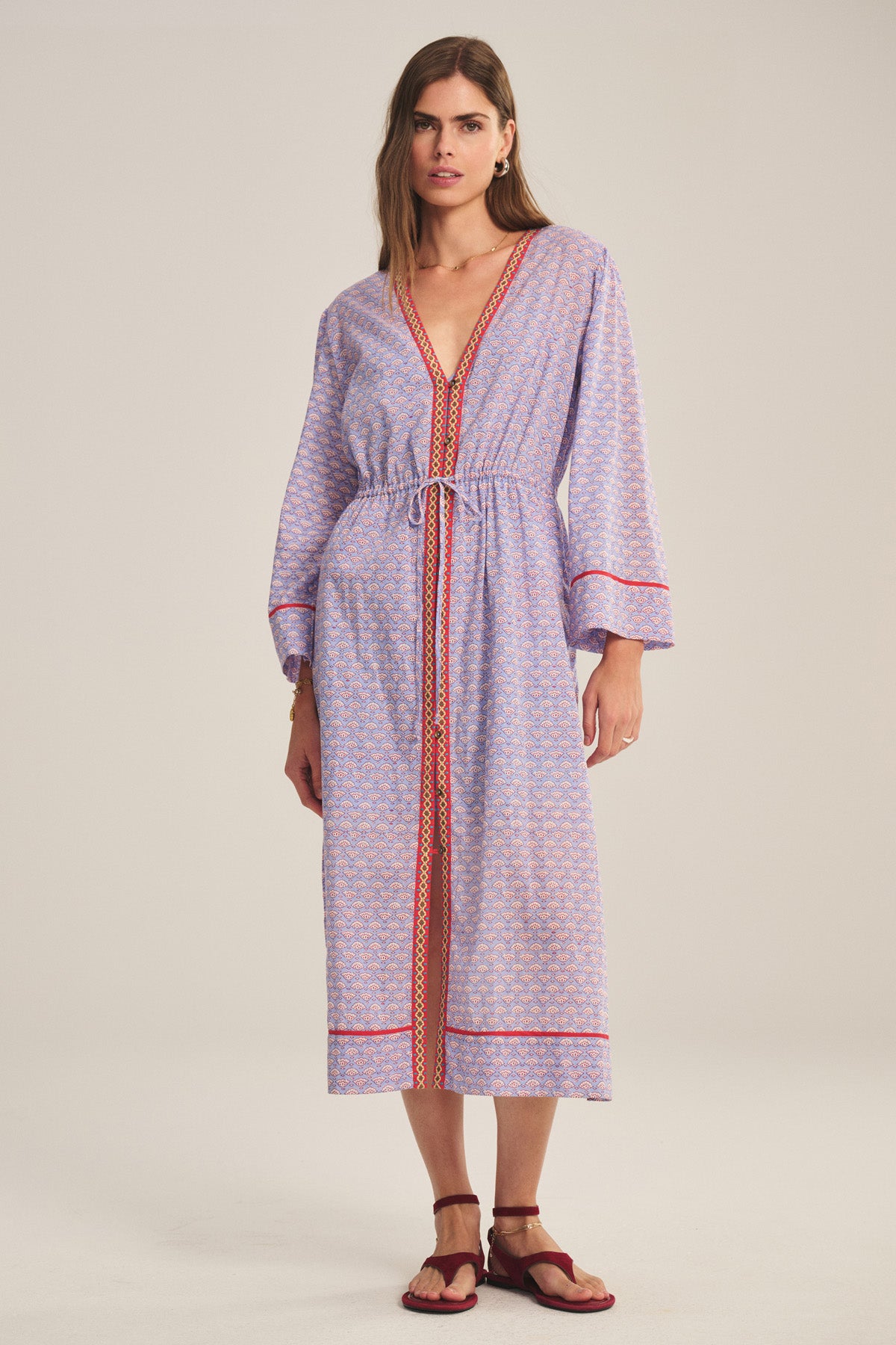 A woman wears the WRENLEY DRESS by Velvet by Graham & Spencer, a light purple cotton voile midi dress with a pattern, red trim, embroidery, long sleeves, and a V-neck. She pairs it with red sandals against a plain background.-40808771420353