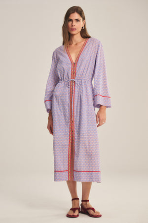 A woman wears the WRENLEY DRESS by Velvet by Graham & Spencer, a light purple cotton voile midi dress with a pattern, red trim, embroidery, long sleeves, and a V-neck. She pairs it with red sandals against a plain background.