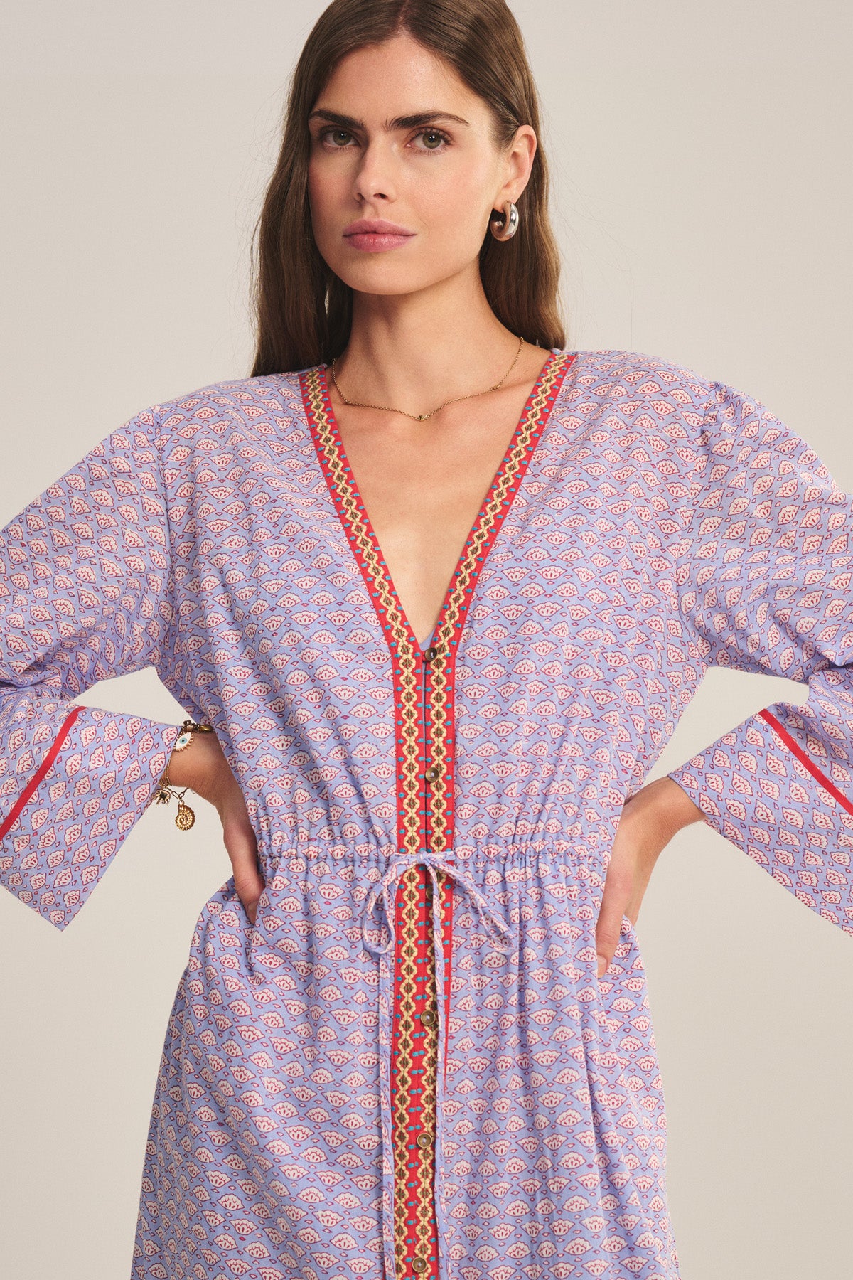 A woman models the WRENLEY DRESS by Velvet by Graham & Spencer, featuring a patterned design, long sleeves, V-neckline, red trim accents, and delicate embroidered details against a plain background.