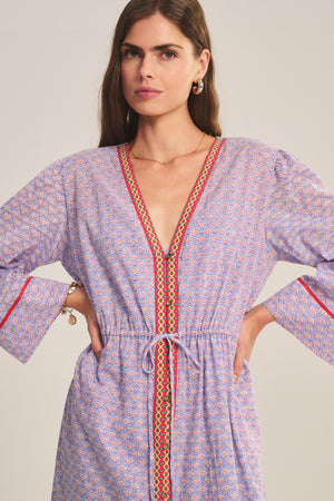 A woman models the WRENLEY DRESS by Velvet by Graham & Spencer, featuring a patterned design, long sleeves, V-neckline, red trim accents, and delicate embroidered details against a plain background.